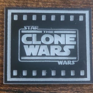 May include: A rectangular black and white patch featuring the Star Wars The Clone Wars logo. The design includes a film strip border with square perforations along the top and bottom edges, framing the bold white text centered on a black background.