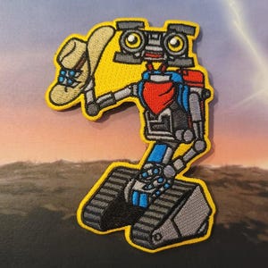 May include: A gray and blue robot wearing a cowboy hat and bandana. The robot is standing on a set of tracks. The robot is a cartoon character.