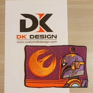 May include: An embroidered patch featuring a purple and orange Mandalorian helmet with a yellow and orange symbol on a purple background. The symbol is a crescent moon with a stylized bird inside. The text "DK DESIGN" and "www.customdkdesign.com" is visible in the background.