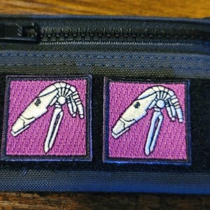 May include: A black zippered pouch with two pink patches featuring white embroidered images of a sci-fi creature.