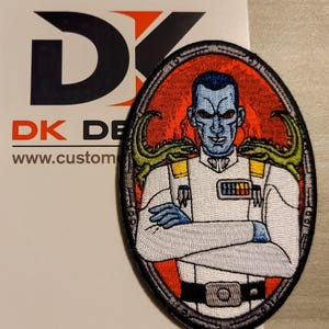 May include: An embroidered patch featuring a blue-skinned character with a stern expression, wearing a white uniform with yellow and red accents. The character is flanked by two green reptilian creatures. The patch is oval-shaped and has a black border.