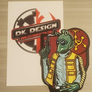 May include: An embroidered patch featuring a green alien character holding a blaster, wearing a yellow vest and a blue shirt. The background is red with a gold symbol.