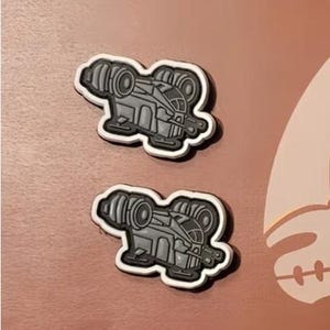 May include: Two gray and white enamel pins featuring a cartoon spaceship with a rounded body and two side engines. The pins are on a brown surface.