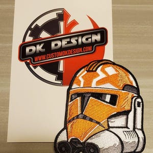 以下が含まれることがあります： スターウォーズのクローン・トルーパーのヘルメットが刺繍されたオレンジと白のワッペン。ワッペンには「DK DESIGN」の文字とウェブサイト「www.customdkdesign.com」が記載されています。
