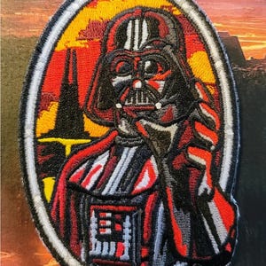 May include: Embroidered patch of Darth Vader from Star Wars. The patch is in an oval shape with a black, red, and white colour scheme. The background depicts a sunset scene with a dark silhouette.