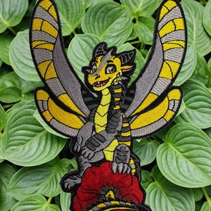 May include: Embroidered patch of a cartoon dragon with yellow and grey wings, a yellow and black body, and a red flower. The dragon sits on the flower, against a backdrop of green leaves. The patch is designed for clothing.