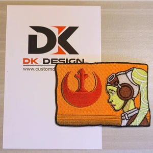 Könnte beinhalten: Ein gestickter Aufnäher mit einem Symbol der Rebellenallianz und einem Porträt einer weiblichen Figur mit grüner Haut und braunen Haaren. Der Aufnäher ist orange mit roten und braunen Akzenten.