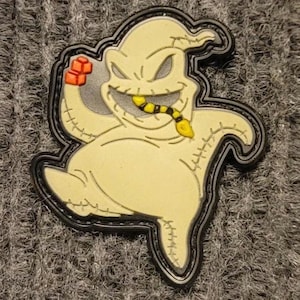 May include: A black and white embroidered patch of a cartoon ghost with a stitched mouth and a yellow worm coming out of its mouth. The ghost is wearing a black outline.