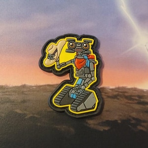 Puede incluir: Un parche bordado amarillo y negro con un robot de dibujos animados que lleva un sombrero de vaquero y un pañuelo rojo. El robot está de pie sobre un conjunto de orugas.