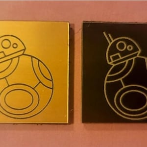 May include: Two square tiles with a black outline of the Star Wars character BB-8. One tile is gold and the other is black.