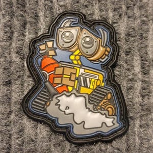 Può includere: Un robot nero e giallo con un corpo blu e un estintore rosso. Il robot ha grandi occhi rotondi e un'espressione amichevole. Il robot è in piedi su una superficie grigia e bianca.