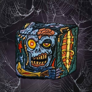 May include: Embroidered patch featuring a zombie design with a blue face, exposed brain, and yellow eye. The patch has a rectangular shape with a green and yellow border, set against a dark background with a spiderweb design.