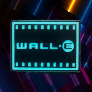Puede incluir: Un parche rectangular con fondo negro, un borde cian brillante y el texto WALL-E en el centro. El diseño del borde imita una tira de película con perforaciones cuadradas a lo largo de los bordes superior e inferior del marco rectangular.