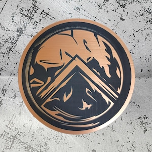 Puede incluir: Un emblema metálico redondo de color cobre con un borde negro. El emblema presenta un diseño estilizado de cordillera en negro, sobre un fondo cobrizo. El diseño es circular y parece ser un elemento decorativo.
