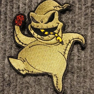 May include: An embroidered patch featuring a cartoon character with a wide grin, holding a red object in its right hand. The character is green and black, with a stitched-on appearance and is wearing a green hat.