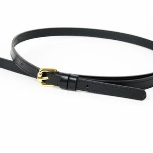 Puede incluir: Una correa de bolso de cuero negro con una hebilla y un cierre dorados. La correa es delgada y parece ser ajustable. La hebilla es rectangular y el cierre es tipo mosquetón.