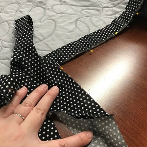 May include: A black and white polka dot fabric is pinned to a gray quilted fabric with yellow pins. The fabric is being sewn, possibly for a quilt or other textile project. The background is a gray quilted fabric.