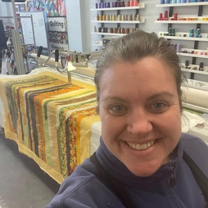 以下が含まれることがあります： 女性がロングアームキルトマシン أمامに立って笑顔を見せています。マシンは、カラフルな糸巻きが置かれた棚のある部屋に設置されています。マシンでキルトがキルティングされています。