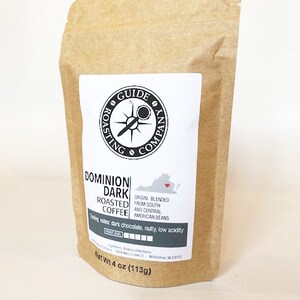 May include: A brown paper bag with a white label. The label has a black compass logo and the text "GUIDE ROASTING COMPANY". The bag contains Dominion Dark Roasted Coffee, which is blended from South and Central American beans. The label also includes tasting notes: dark chocolate, nutty, low acidity. The bag is labeled with a net weight of 4 ounces (113g).