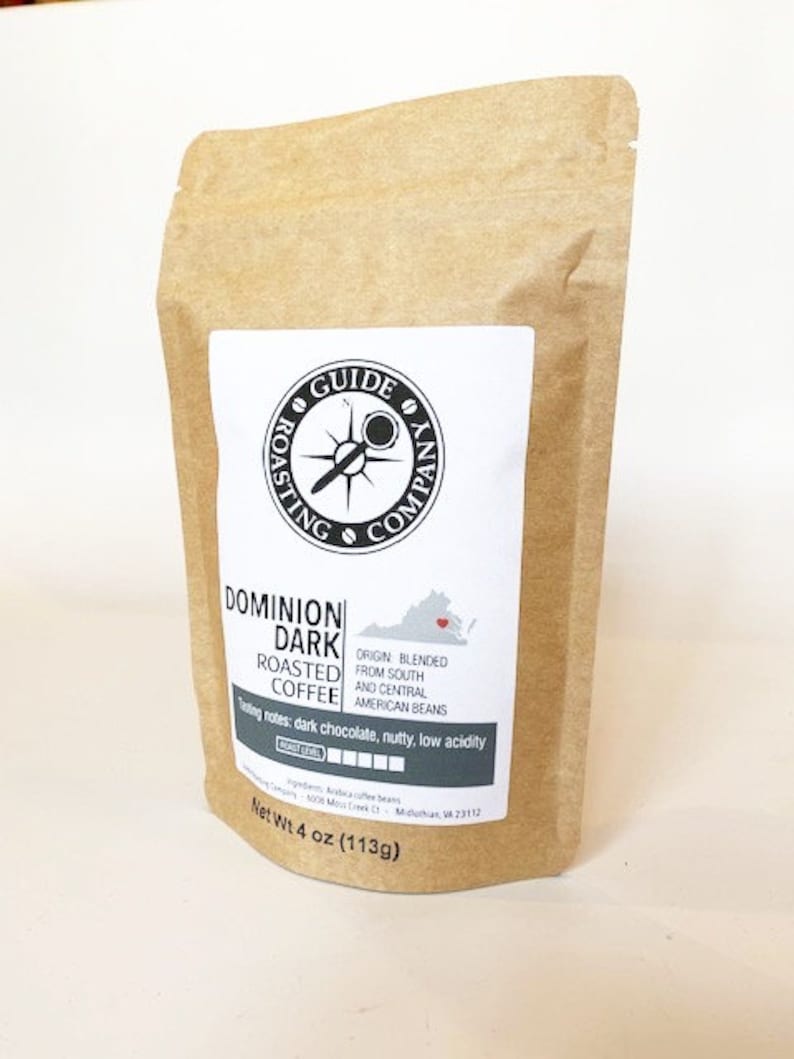 May include: A brown paper bag with a white label. The label has a black compass logo and the text "GUIDE ROASTING COMPANY". The bag contains Dominion Dark Roasted Coffee, which is blended from South and Central American beans. The label also includes tasting notes: dark chocolate, nutty, low acidity. The bag is labeled with a net weight of 4 ounces (113g).