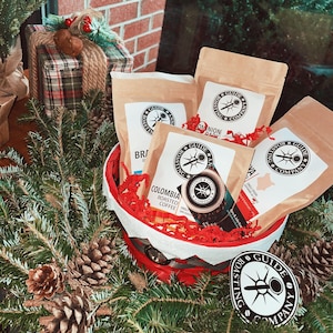 May include: A red and white basket filled with four bags of coffee beans. The bags are labeled with the text "Millston Guide & Roast Co." and different coffee blends. The basket is surrounded by pine branches and pine cones.