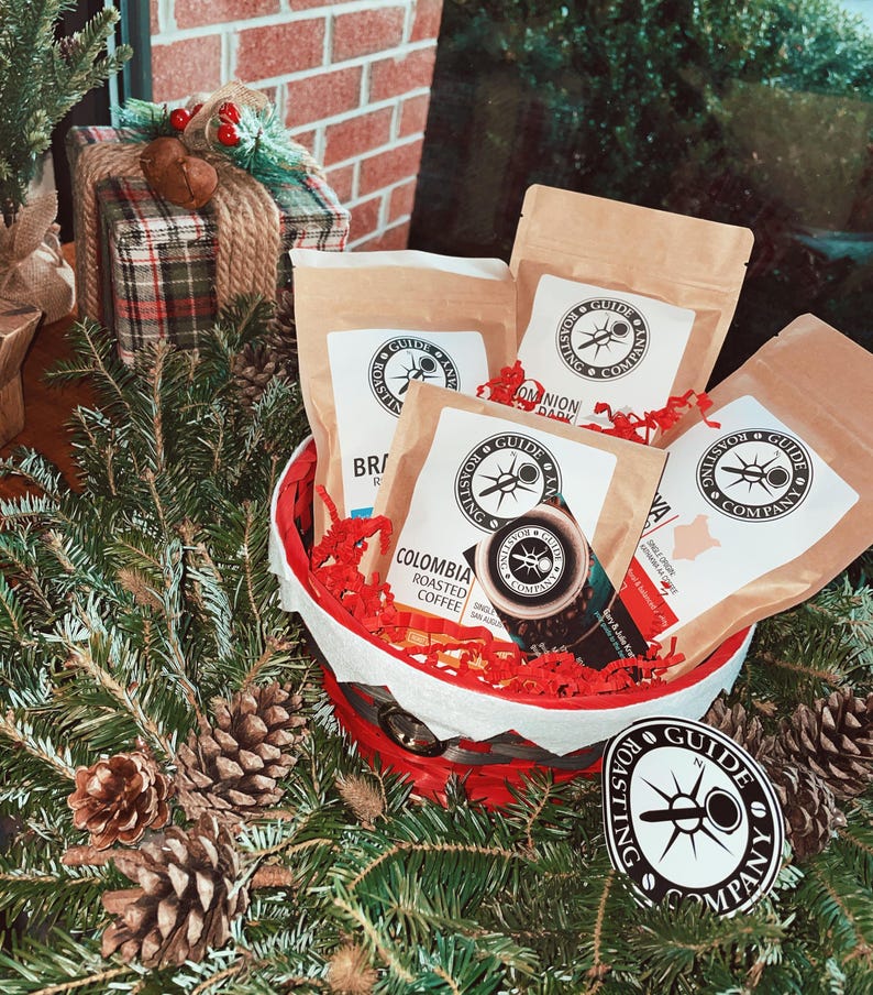 May include: A red and white basket filled with four bags of coffee beans. The bags are labeled with the text "Millston Guide & Roast Co." and different coffee blends. The basket is surrounded by pine branches and pine cones.