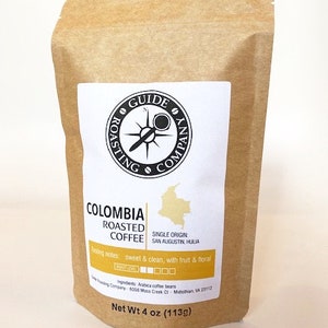 May include: A brown paper bag with a label that says "Colombia Roasted Coffee" with a compass logo and the text "Guide Roasting Company". The label also includes the text "Single Origin: San Agustin, Huila" and "Tasting notes: sweet & clean, with fruit & floral". The bag is labeled "Net Wt 4 oz (113g)".