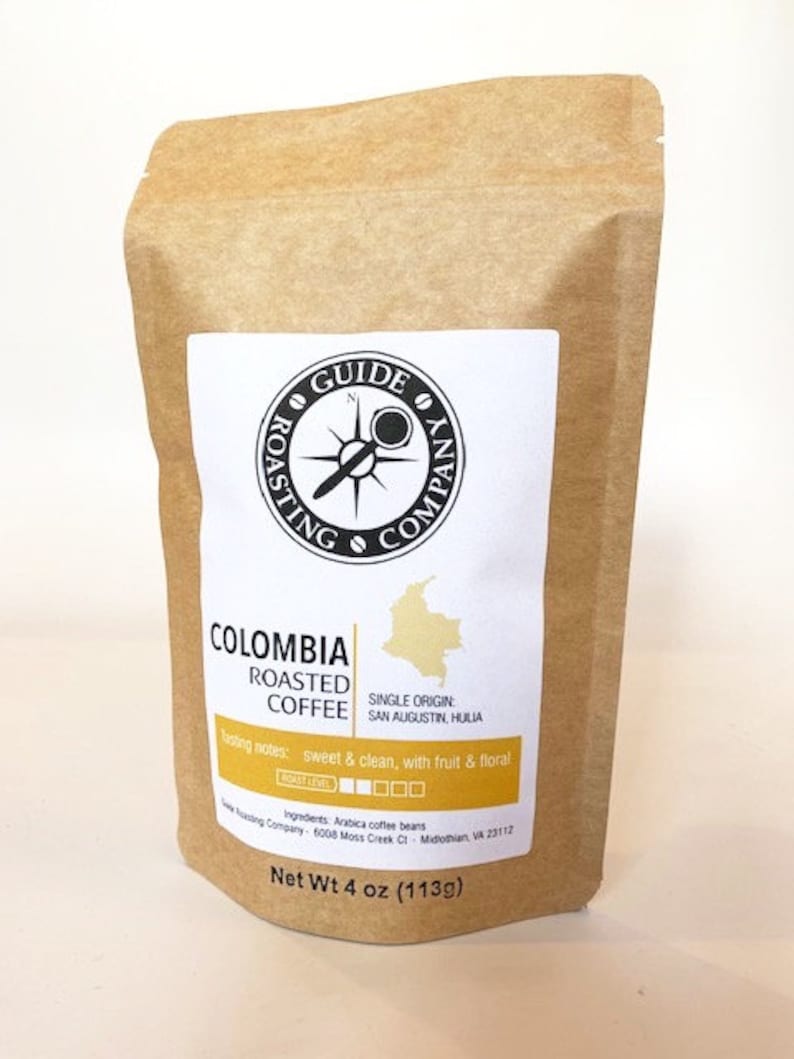 May include: A brown paper bag with a label that says "Colombia Roasted Coffee" with a compass logo and the text "Guide Roasting Company". The label also includes the text "Single Origin: San Agustin, Huila" and "Tasting notes: sweet & clean, with fruit & floral". The bag is labeled "Net Wt 4 oz (113g)".