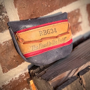 Könnte beinhalten: Ein handgefertigter Golfschlägerkopfschutz aus dunkelblauem Leder. Er hat einen beigefarbenen Lederaufnäher mit dem Text "RBG34" und "The Finest In The Field!" in braunen Buchstaben, mit roten Nähten.
