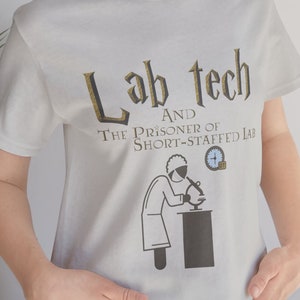 May include: A light gray t-shirt with a gold graphic that says "Lab Tech and the Prisoner of Short-Staffed Lab". The graphic also includes a cartoon of a person wearing a lab coat and using a microscope.