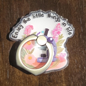 May include: A clear phone ring holder with a silver ring. The holder has a floral design with a microscope and the text "Enjoy the little things in life".