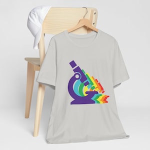 May include: A light gray t-shirt with a rainbow-colored microscope graphic. The microscope is purple with a rainbow-colored lens and a rainbow-colored base.