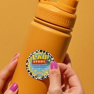May include: Mustard-yellow water bottle with a matching cap and handle. A round sticker with the text "LAB STORY" and a cow-print border is being applied to the bottle. The background is a solid orange.