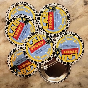 May include: A collection of round buttons with a black and white cow-print border. Each button features the words "LAB STORY" in red and yellow, with the text "Lab Week 2026" and "To Infinity and Beyond for our Patients!"