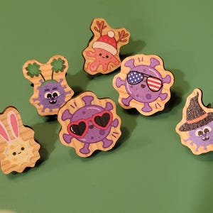 May include: A collection of wooden pins with cartoon virus designs. Each pin features a different festive theme, including a bunny, shamrocks, a Santa hat, heart-shaped sunglasses, American flag sunglasses, and a witch hat. The pins are on a green background.