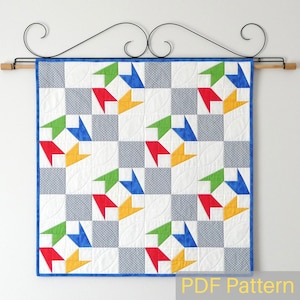 Könnte beinhalten: Ein farbenfroher Quilt mit einem geometrischen Muster aus blauen, grünen, roten und gelben Dreiecken. Der Quilt hängt an einer Wand mit einer Holzstange und schwarzen Metallhaken. Der Quilt ist aus weißem Stoff mit einem grau-weißen Tupfenmuster gefertigt. Der Quilt ist mit einem blauen Besatz eingefasst. Der Text "PDF Pattern" ist in Gelb am unteren Rand des Bildes gedruckt.