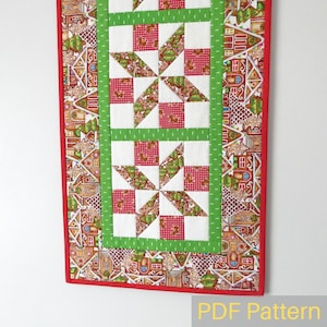 May include: A red, green and white patchwork table runner with a gingerbread house pattern border. The runner is made of fabric squares and features a starburst design. The text "PDF Pattern" is visible at the bottom of the image.
