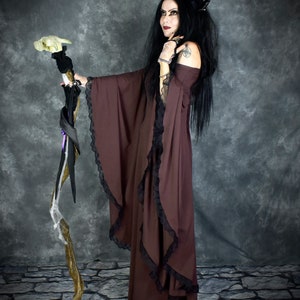 Pu&ograve; includere: Una donna con un vestito color borgogna con bordatura in pizzo nero e una fascia nera con corna tiene un bastone di legno con un teschio bianco in cima. Il bastone &egrave; avvolto in tessuto nero e ha un motivo a ragnatela bianca.
