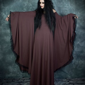 Pu&ograve; includere: Una donna indossa un lungo abito maxi marrone scuro con maniche lunghe e bordi in pizzo. L'abito ha una silhouette fluida e drammatica. Indossa una fascia per capelli nera con un motivo floreale e ha i capelli lunghi e scuri.