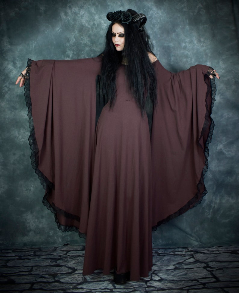 Pu&ograve; includere: Una donna indossa un lungo abito maxi marrone scuro con maniche lunghe e bordi in pizzo. L'abito ha una silhouette fluida e drammatica. Indossa una fascia per capelli nera con un motivo floreale e ha i capelli lunghi e scuri.