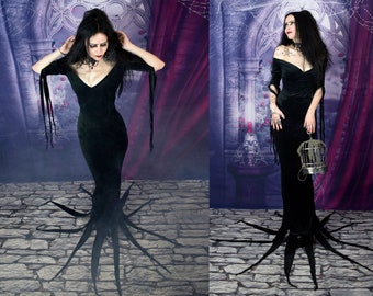 May include: A floor-length black velvet dress featuring a V-neckline, long sleeves with hanging fabric strips, and a hemline adorned with multiple long, pointed fabric extensions that spread outward across the floor in a star-like pattern.