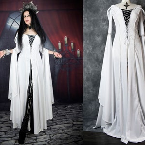 Puede incluir: Un vestido de novia gótico blanco con un corpiño con cordones y mangas largas. El vestido tiene una falda fluida y una cola. El vestido lo lleva una modelo con el pelo largo y negro y una corona.