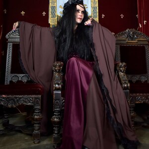 Pu&ograve; includere: Una donna vestita con una lunga tunica marrone scuro con bordi in pizzo nero siede su un trono. Ha i capelli lunghi e neri e indossa una gonna viola scuro. Il trono &egrave; intagliato in modo elaborato e ha un cuscino di velluto rosso.