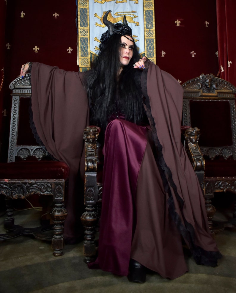 Pu&ograve; includere: Una donna vestita con una lunga tunica marrone scuro con bordi in pizzo nero siede su un trono. Ha i capelli lunghi e neri e indossa una gonna viola scuro. Il trono &egrave; intagliato in modo elaborato e ha un cuscino di velluto rosso.