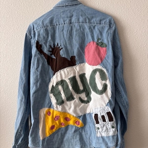 May include: Light blue denim shirt with NYC-themed patches. Features a brown Statue of Liberty, a pink apple, a yellow pizza slice, and a white patch with "nyc" in green letters. A bus patch is also visible.