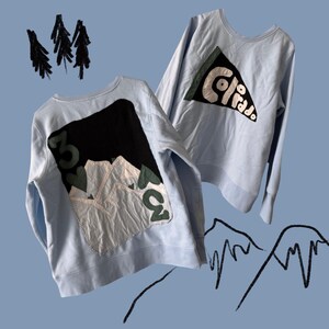 May include: Two pale blue sweatshirts with mountain-themed patches. One has a black mountain design with the number "303" and a white and green mountain graphic. The other has a triangle patch with the word "Colorado".