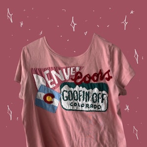 May include: A pale pink t-shirt with a scoop neck and short sleeves. The shirt has embroidered patches with the words "Denver Coors" and "Goofin Off Colorado". A Colorado flag patch is also visible.