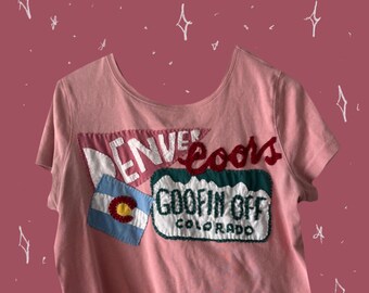 May include: A pale pink t-shirt with a scoop neck and short sleeves. The shirt has embroidered patches with the words "Denver Coors" and "Goofin Off Colorado". A Colorado flag patch is also visible.