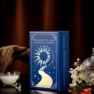 Pode incluir: Uma caixa azul com detalhes dourados em folha apresenta um baralho de tarot chamado "The Golden Path Tarot". A caixa tem um design de sol e lua com um caminho dourado que leva &agrave; lua. O texto na caixa diz "A 78-Card Deck and Guidebook" e "Blue Moon Edition".