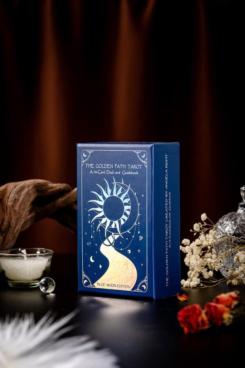 Pode incluir: Uma caixa azul com detalhes dourados em folha apresenta um baralho de tarot chamado "The Golden Path Tarot". A caixa tem um design de sol e lua com um caminho dourado que leva &agrave; lua. O texto na caixa diz "A 78-Card Deck and Guidebook" e "Blue Moon Edition".